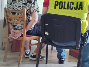 zatrzymany podczas przedstawiania mu zarzutów siedzi na krześle z tyłu na rekach nałożone kajdanki, obok pilnuje go policjant