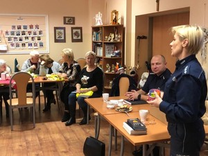 Foto przedstawia policjantów oraz seniorów biorących udział w debacie społecznej. Podczas spotkania rozdawane są również  upominki dla seniorów które mają wpłynąć na poprawę poczucia bezpieczeństwa.