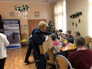 Foto przedstawia policjantów oraz seniorów biorących udział w debacie społecznej. Podczas spotkania rozdawane są również  upominki dla seniorów które mają wpłynąć na poprawę poczucia bezpieczeństwa.